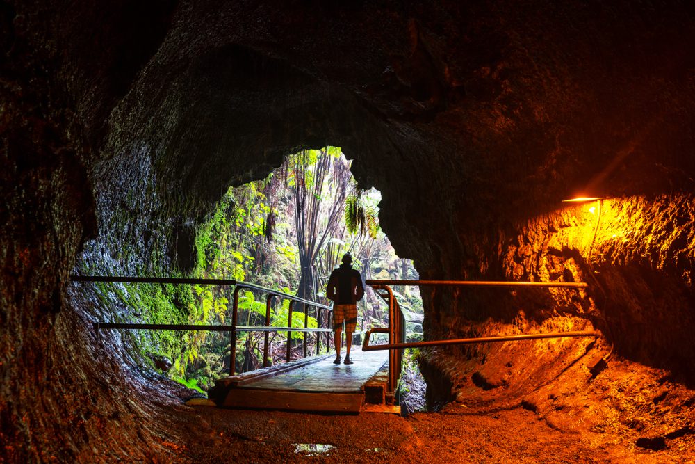 Silhouette of person in lava tube walking out, sun and trees in backgroung outside.