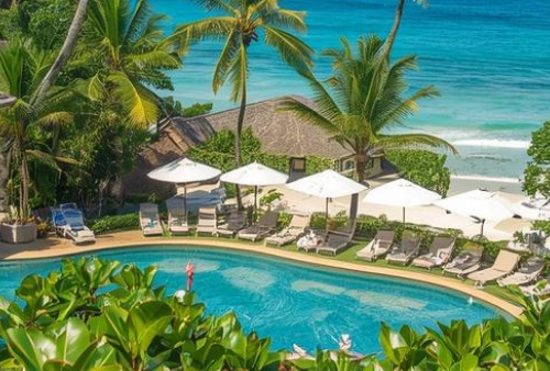 Tropical beach hotel with pool and foliage, next to beach.