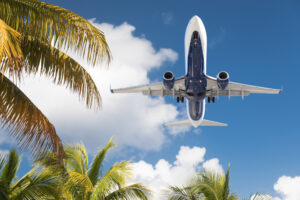 Jet airplane flying overhead with palm trees, blue sky and white clouds.