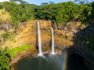 Twin waterfalls cascade from a rocky cliff into a pool, surrounded by lush greenery with a rainbow at the base.