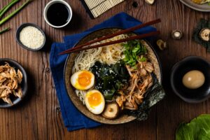 Traditional ramen with jerked pork or chicken. With udon or ramen noodles. Served in classic bowls. Gyoza dumplings and mushrooms in the background. Natural wooden background.